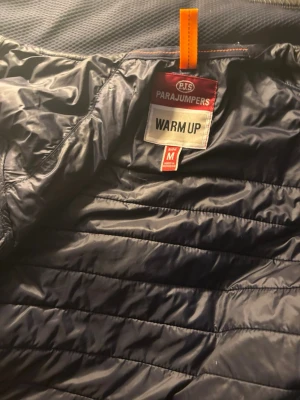 Mörkblå dunväst Parajumpers M - Snygg mörkblå dunväst från Parajumpers med gul detalj vid dragkedjan och orange hängare. Västen har quiltad design och dragkedja framtill. Perfekt för lager-på-lager och håller dig varm under kyliga dagar.