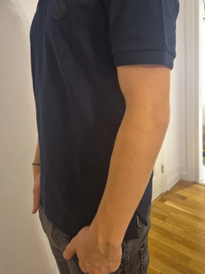 Mörkblå pikétröja från Moncler - Klassisk mörkblå pikétröja från Moncler med korta ärmar och krage. Tröjan har knappslå framtill och en diskret Moncler-logga på bröstet. Perfekt för en stilren och avslappnad look.