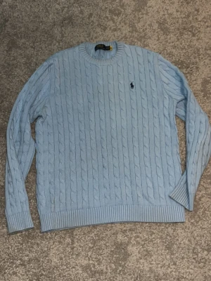 Ljusblå kabelstickad tröja Polo Ralph Lauren - Snygg ljusblå kabelstickad tröja från Polo Ralph Lauren med klassisk rund halsringning och broderad logga på bröstet. Tröjan har ribbade muddar vid ärmslut och nederkant. Perfekt för dig som gillar preppy stil och mjuka material.