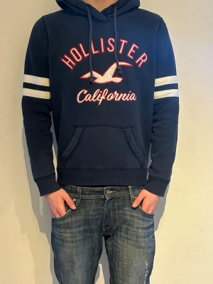 Marinblå vintage hoodie från Hollister - Snygg marinblå hoodie från Hollister med vit och röd brodyr på bröstet där det står 'Hollister California' och en mås. Två vita ränder på ärmarna och klassisk känguruficka framtill. Huva med dragsko och ribbade muddar. Perfekt för en avslappnad stil.