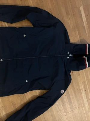 Marinblå vindjacka från Moncler - Snygg marinblå vindjacka från Moncler med dragkedja framtill, två sidofickor med tryckknappar och Moncler-logga på ärmen. Jackan har ribbade muddar med rödvita detaljer vid ärmslutet. Perfekt för dig som vill ha en stilren och sportig look.