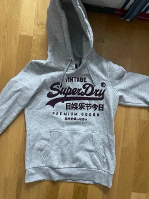 Grå Superdry hoodie med tryck -  sopig grå hoodie från Superdry med stor vinröd logga och japansk text på bröstet. Klassisk känguruficka framtill, dragsko i huvan och mjukt material som känns skönt mot huden. Perfekt för chill dagar eller när du vill ha en avslappnad stil.