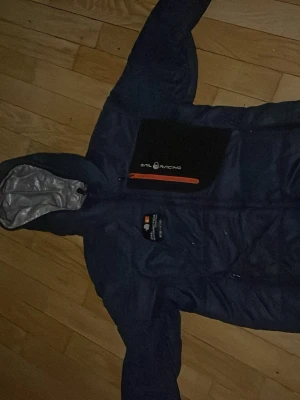 Mörkblå pufferjacka Sail Racing - Snygg mörkblå pufferjacka från Sail Racing med luva och dragkedja framtill. Jackan har en sportig look och är tillverkad i slitstarkt syntetmaterial. Perfekt för kyliga dagar och har en klassisk passform.