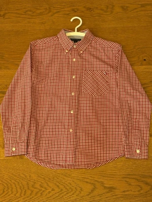 Rutig röd skjorta från Tommy Hilfiger - Klassisk röd- och vitrutig skjorta från Tommy Hilfiger med lång ärm, knappar framtill och bröstficka med liten logga. Skjortan har en normal passform och är tillverkad i mjuk bomull. Perfekt för en avslappnad men snygg stil. Den passar ca 158. Mer bilder kan fås om det önskas. Använd Max 2-3 ggr