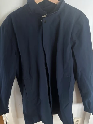 Mörkblå rock från Lindbergh - Stilren mörkblå rock från Lindbergh med klassisk krage och dold knäppning framtill. Rocken har raka fickor på sidorna och är tillverkad i ett slätt, vattenavvisande material. Perfekt för dig som gillar en clean och modern look.nypris 3350!