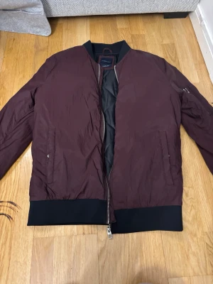 Vinröd bomberjacka från Arket - Snygg vinröd bomberjacka från Arket med svarta muddar och dragkedja framtill. Jackan har fickor på sidorna och en klassisk ärmficka med dragkedja. Materialet är glansigt och lätt, perfekt för en trendig streetwear-look.