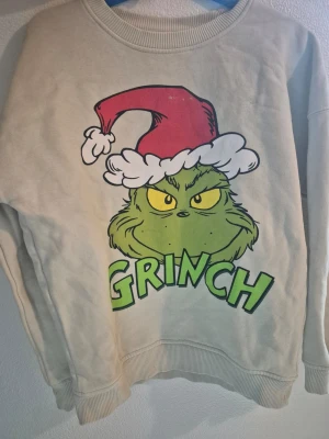 Barn Beige stickad tröja med Grinchen  stl 134 - Cool beige stickad tröja med stort tryck av Grinchen i grönt och gult, samt röd tomteluva och texten 'GRINCH' framtill. Tröjan har rund halsringning och ribbade muddar vid ärmar och nederkant. Perfekt för dig som gillar jul och popkultur.