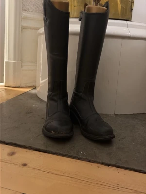 Svarta höga ridstövlar i läder - Säljer ett par klassiska svarta ridstövlar i äkta läder. De har en stilren, hög modell med rundad tå och diskreta sömmar. Perfekta för dig som gillar en tidlös look till ridningen eller vill ha ett par boots med attityd.