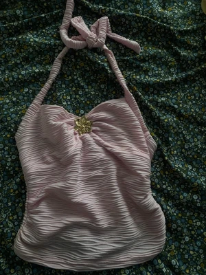 Ljusrosa ribbad halterneck topp - Supersöt ljusrosa halterneck topp med ribbad struktur och en guldfärgad blomdetalj framtill. Toppen knyts i nacken och har en mjuk, stretchig känsla. Perfekt för dig som vill ha en feminin och trendig look.