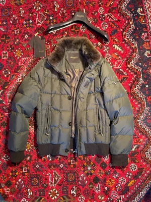 MooRER - Stilren grön pufferjacka från MooRER med avtagbar kaninpäls på kragen, ribbade muddar i kashmir blandning och dragkedjor på fickorna. Jackan har quiltad design och detaljer i svart, perfekt för kalla vinterdagar. Snygg och funktionell med exklusiv känsla. Storlek 50 passar som en M. Nypris runt 20000kr. Zippen kan va lite små bråkig men man vänjer sig efter enstaka gånger. 