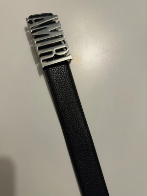 Svart skärp från Amiri med silverdetalj - Snyggt svart bälte från Amiri med en stor, silverfärgad metallspänne där märkesnamnet står i 3D. Bältet är gjort i skinn med en stilren och modern look som passar till jeans eller byxor.
