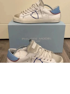 Philippe Model vita sneakers blå detalj - Snygga vita sneakers från Philippe Model med blå hälkappa och röda detaljer. Skorna har beige mocka framtill, vita skosnören och en cool blå logga på sidan. Yttersulan är i gummi med fiskbensmönster. Perfekta för en trendig och avslappnad look.