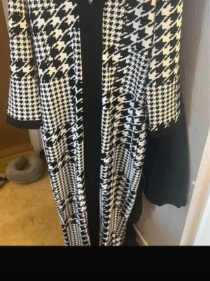 Svartvit rutig långklänning - Säljer en svartvit långklänning med klassiskt rutigt mönster och trekvartsärm. Klänningen har en markerad svart kant framtill och är i ett mjukt, stickat material. Perfekt för dig som gillar statement-plagg med retrovibbar.