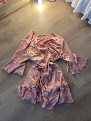 Rosa satinmorgonrock med volangärm - Snygg och mjuk morgonrock i glansigt rosa satintyg. Modellen har vida volangärmar och knytskärp i midjan för en avslappnad och chic look. Perfekt för att chilla hemma i stil.