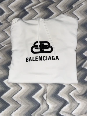 Balenciaga Logo Hoodie - Snygg vit hoodie från Balenciaga med svart tryckt logga på bröstet. Klassisk modell med huva och dragsnören. Perfekt för dig som gillar streetwear och vill sticka ut med en stilren look.