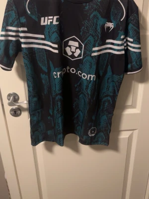 UFC x Venum svart/grön sporttröja - Cool svart och grön sporttröja från Venum i samarbete med UFC. Tröjan har ett unikt ormmönster, tryckta loggor från UFC och Crypto.com samt vita ränder på ärmarna. Perfekt för träning eller som supportertröja. Tillverkad i lätt och snabbtorkande material. OBS de känns som medium eftersom de är compressed.