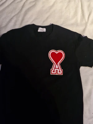 Svart Ami t-shirt med röd hjärtlogga - Säljer en svart t-shirt från Ami med deras ikoniska röda hjärta och A-logga i frotté på bröstet. T-shirten har rund hals och är tillverkad i mjukt bomullsmaterial. Perfekt för dig som gillar streetwear och vill ha något som sticker ut.