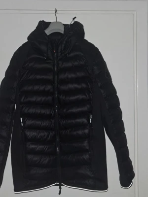 Svart dunjacka från Zavetti Canada - Snygg svart dunjacka från Zavetti Canada med quiltad design och huva. Jackan har dragkedja framtill, två sidofickor med dragkedja och en patch med logga på ärmen. Perfekt för kalla vinterdagar och riktigt stilren look.