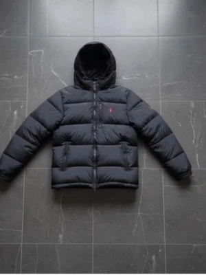 Svart dunjacka från Polo Ralph Lauren - Svart dunjacka från Polo Ralph Lauren med huva och dragkedja framtill. Jackan har två fickor med dragkedja och en liten röd logga på bröstet. Klassisk puffer-modell som håller dig varm under kalla dagar.