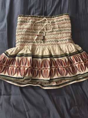 Beige kortkjol med mönster från Zara - Supersöt beige kortkjol från Zara med elastisk midja och snörning med pärldetaljer. Kjolen har ett unikt brunt och rött mönster längs fållen och volangkant som ger en bohemisk vibe. Perfekt för varma dagar och sommarens alla äventyr!