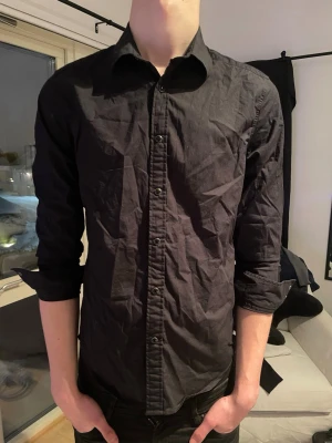 Svart långärmad skjorta - Säljer en svart långärmad skjorta med klassisk krage och knäppning framtill. Skjortan har en slim fit-passform och är tillverkad i ett tunt, lätt material som känns bekvämt mot huden. Perfekt att styla med jeans eller chinos för en clean look.