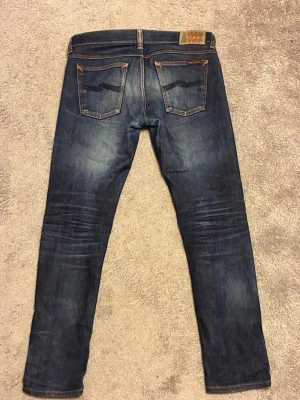Nudie jeans vintage - Säljer ett par mörkblå jeans från Nudie med klassiska kontrastsömmar och slitningar framtill. Jeansen har fem fickor och är tillverkade i stretchig bomullsdenim för en bekväm passform.