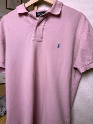 Ljusrosa pikétröja från Polo Ralph Lauren - Ljusrosa pikétröja från Polo Ralph Lauren med klassisk krage, korta ärmar och blå broderad logga på bröstet. Tillverkad i bomull med pikéstruktur och två knappar vid halsen. Perfekt för en avslappnad men ändå stilren look.