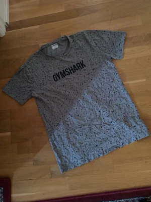 Gymshark t-shirt - Snygg grå t-shirt från Gymshark med svart logga på bröstet. T-shirten har korta ärmar och ett unikt prickigt mönster över hela tyget. Perfekt för dig som gillar sportig och trendig stil. Storlek: S