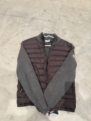  Moncler cardigan grå - Snygg cardiganjacka från Moncler med quiltad, mörkgrå front och stickade ärmar och rygg i ljusare grått. Jackan har dragkedja framtill, två sidofickor med dragkedja och Moncler-logga på ärmen. Perfekt för lager-på-lager-stil.