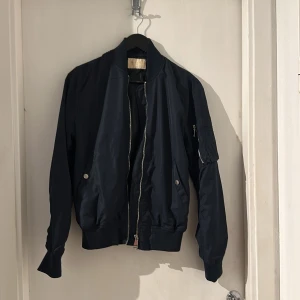 Marinblå  bomberjacka  - Snygg svart bomberjacka med klassisk siluett, ribbade muddar och silverfärgad dragkedja. Jackan har fickor med tryckknappar och en extra ficka med dragkedja på ärmen. Perfekt för dig som gillar en stilren och urban look.