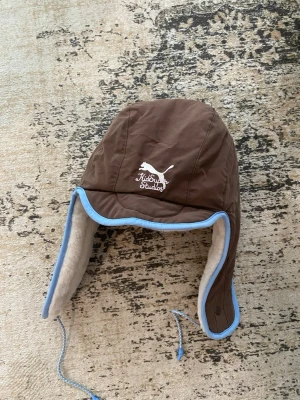 Brun trappermössa från KidSuper Studios - Säljer en brun trappermössa från KidSuper Studios med ljusblå kantband och vitt, mjukt foder. Mössan har öronlappar och knytband och är reversible. Jag har tyvärr klippt av lapparna men den är köpt från BSTN store.