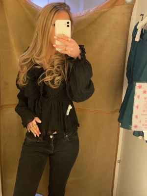 Svart plisserad blus med volanger - Trendig svart blus med plisserat tyg och volangdetaljer framtill. Blusen har v-ringning och långa ärmar med resår vid ärmslut. Perfekt för dig som gillar en chic och stilren look. Materialet känns lätt och luftigt.