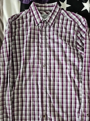 Rutig skjorta från DKNY i lila och vit - Snygg rutig skjorta från DKNY i lila och vit med klassisk krage och knappar framtill. Skjortan har långa ärmar med manschett och är tillverkad i mjuk bomull. Perfekt för dig som gillar stilrena och färgglada plagg.