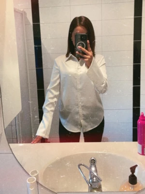 Vit satinliknande skjorta - Stilren vit skjorta i satinliknande material med klassisk krage och långa ärmar. Skjortan har en rak passform och knäppning framtill. Perfekt för dig som gillar en clean och enkel look.