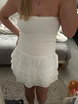Vit axelbandslös kortklänning - Söt vit axelbandslös kortklänning med smockad överdel och volangig kjol. Klänningen har broderade små blommor och är perfekt för varma dagar. Lätt och luftig känsla, passar dig som gillar en feminin och somrig stil. Perfekt för student eller midsommar 