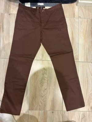 Brun röda chinos från gant 34/32 - Snygga bruna chinos från Gant i klassisk rak modell. Tillverkade i ett mjukt och slitstarkt bomullstyg med bälteshällor och knappgylf. Perfekta för dig som vill ha en stilren och enkel look.