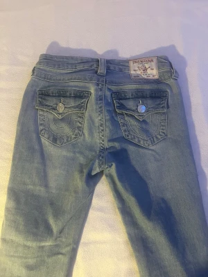 Ljusblå True Religion jeans - Säljer ett par ljusblå jeans från True Religion med klassiska kontrastsömmar och snygga fickdetaljer bak. Jeansen har raka ben, fem fickor och silverfärgade knappar. Perfekta för en avslappnad och trendig look.