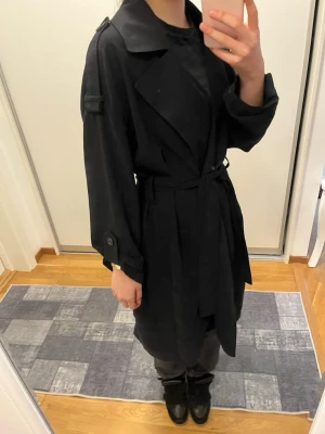 Svart trenchcoat från Vero Moda - Stilren svart trenchcoat från Vero Moda med klassisk krage, axelklaffar och knytskärp i midjan. Jackan har långa ärmar med knappdetaljer och är tillverkad i ett mjukt tyg som ger en snygg siluett. Perfekt för dig som gillar minimalistisk och tidlös stil.