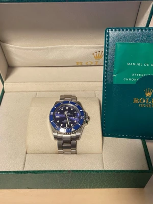 Blå och silver klocka med låda - Snygg Submariner med blå urtavla och blå vridring. Klockan har ett klassiskt armband i silverfärgat stål och tydliga vita markeringar på urtavlan. Kommer i originalbox med certifikat och garantikort. Perfekt för dig som gillar exklusiva accessoarer. Ett fel är att det är svårt att ändra tiden på klockan och det andra felet är att armbandet ibland öppnar sig.