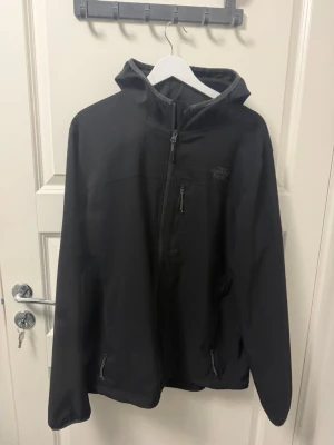 Svart vindjacka från The North Face - Snygg svart vindjacka från The North Face med huva och dragkedja framtill. Jackan har två sidofickor med dragkedja och en bröstficka. Tillverkad i ett lätt och slitstarkt material som skyddar mot vind. Perfekt för blåsiga dagar och outdoor-äventyr.
