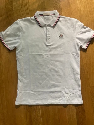 Moncler pike - Säljer en klassisk vit pikétröja från Moncler med krage och knappar framtill. Tröjan har tunna röda och blå ränder på krage och ärmslut samt Moncler-logga på bröstet. Tillverkad i bomull och har en normal passform.