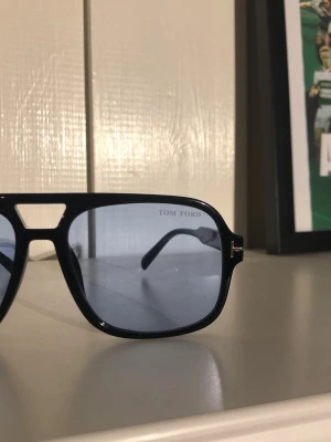 Svarta Tom Ford pilotbågar solglasögon - Snygga svarta solglasögon från Tom Ford med klassisk pilotform och mörktonade glas. Ramen är i blank svart plast med guldfärgade detaljer vid gångjärnen. Perfekta för dig som vill ha en stilren och trendig accessoar.