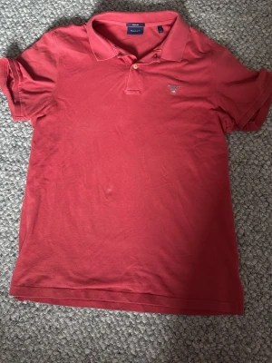 Röd piké från Gant - Klassisk röd pikétröja från Gant med liten broderad logga på bröstet. Tröjan har krage, knappar framtill och korta ärmar. Perfekt för en avslappnad men ändå stilren look.