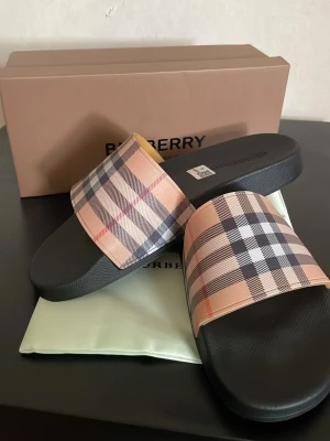 Burberry rutiga tofflor - Säljer ett par snygga tofflor från Burberry med klassiskt beige, svart och rött rutmönster på remmen. Tofflorna har en svart sula i syntetmaterial och är perfekta för att ge din look en lyxig touch. Medföljer originalkartong och dustbag.