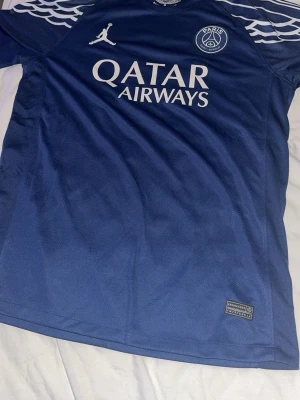 PSG matchtröja blå O.Dembélé #10 - Snygg blå Paris Saint-Germain matchtröja med vita detaljer och tryck. Framsidan har PSG-logga, Jordan-logga och Qatar Airways sponsor. Baksidan har O.Dembélé och nummer 10. Tillverkad i lätt och ventilerande material, perfekt för fotboll eller träning.