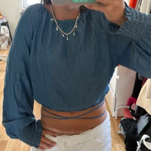 Blå croppad topp med snörning - Trendig blå croppad topp med långa ärmar och smockade muddar. Toppen har snörning i midjan som kan knytas runt kroppen för en snygg detalj. Perfekt till jeans eller kjol för en cool look. Det är storlek M men passar Xs-M då den knyts i midjan. 