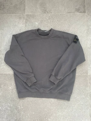 Grå tröja från Stone Island - Säljer en grå sweatshirt från Stone Island med klassisk rund hals och ribbade muddar. Tröjan har den ikoniska Stone Island-patchen på vänster ärm. Materialet är mjuk bomull och passformen är normal, perfekt för chill dagar. Har använt tröjan några få gånger och finns inga hål eller liknande på tröjan, skicket är precis som nytt! Pris går att diskutera☺️