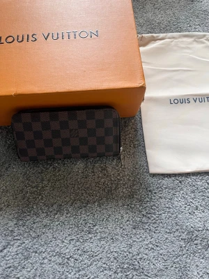 brun plånbok - Snygg brun plånbok. Plånboken har dragkedja runtom och är tillverkad i canvas med detaljer i läder. Perfekt för dig som gillar lyxiga accessoarer och vill ha ordning på kort och kontanter. Ask och dustbag följer ej med.