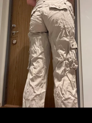 Beige cargobyxor med fickor - Säljer ett par beige cargobyxor med flera stora fickor på sidorna och bak. Byxorna har en relaxed passform och är perfekta för en chill och avslappnad stil. Tillverkade i bomull för skön känsla och andningsförmåga. Passar dig som gillar praktiska detaljer och streetwear. Lite slitningar på snöret på ena midjespännet. Annars i fint skick