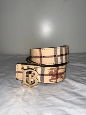 Beige rutigt bälte från Burberry - Snyggt bälte från Burberry i klassiskt beige och brunt rutigt mönster med guldigt B-spänne. Perfekt accessoar för att lyfta din outfit med en lyxig touch. Tillverkat i skinn med ikoniskt Burberry-mönster.